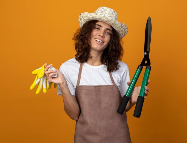 Улыбающаяся молодая женщина-садовник в униформе в садовой шляпе держит ножницы в перчатках
