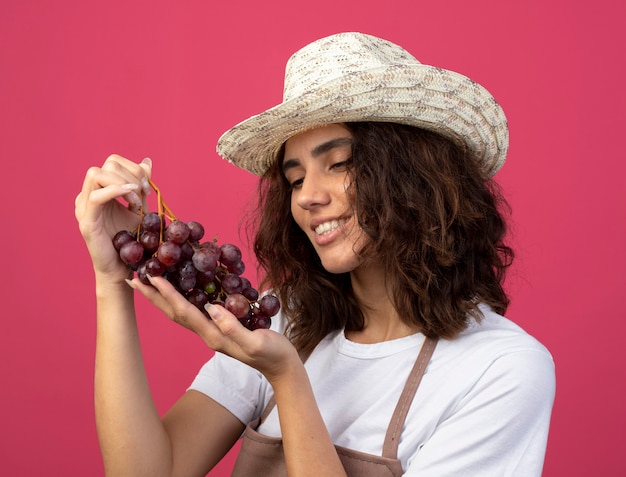 Free photo smiling young female gardener in uniform wearing gardening hat holding and looking at grapes isolated on pink