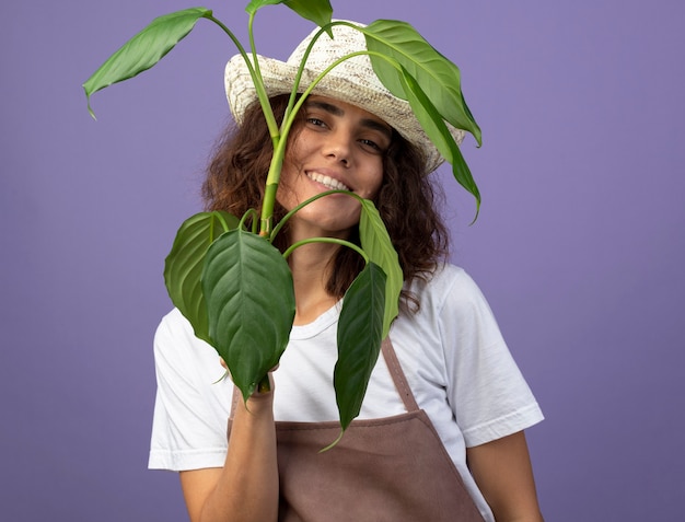 Foto gratuita sorridente giovane giardiniere femminile in uniforme che indossa il cappello da giardinaggio azienda pianta