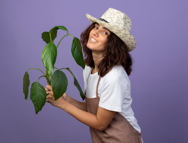 식물을 들고 원예 모자를 쓰고 제복을 입은 젊은 여성 정원사 미소