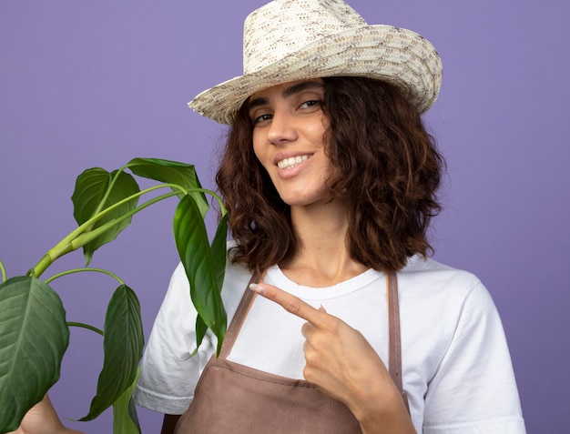 Smiling young female gardener in uniform wearing gardening hat holding and points at plant
