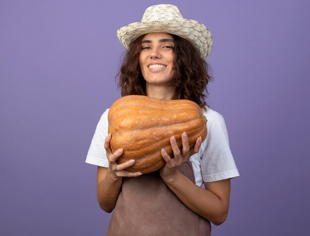 Smiling young female gardener in uniform wearing gardening hat holding pumpkin