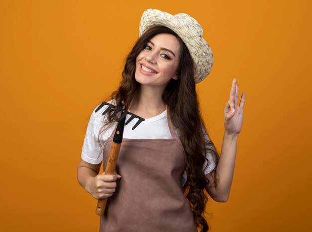 Улыбающаяся молодая женщина-садовник в униформе в садовой шляпе держит грабли и жестикулирует знак рукой на оранжевой стене с копией пространства