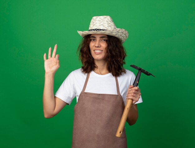 Smiling young female gardener in uniform wearing gardening hat holding rake showing okay gesture