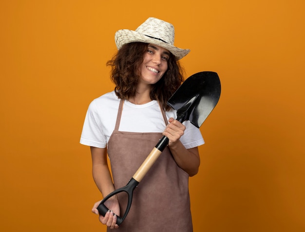 Free photo smiling young female gardener in uniform wearing gardening hat holding spade