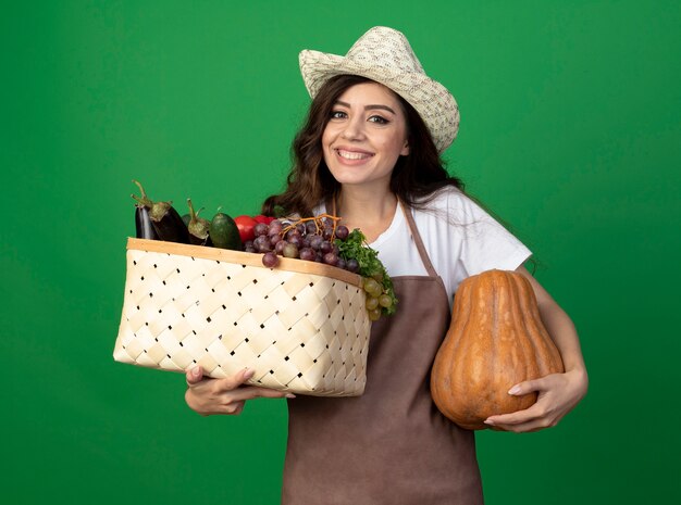 Smiling young female gardener in uniform wearing gardening hat holding vegetable basket and pumpkin isolated on green wall with copy space