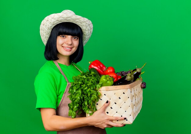 Smiling young female gardener in uniform wearing gardening hat holding vegetable basket