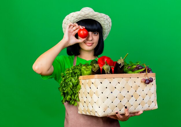 Smiling young female gardener in uniform wearing gardening hat holding vegetable basket