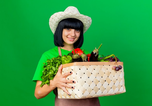 야채 바구니를 들고 원예 모자를 쓰고 유니폼에 웃는 젊은 여성 정원사