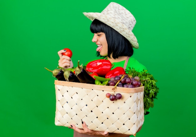 Smiling young female gardener in uniform wearing gardening hat holding vegetable basket