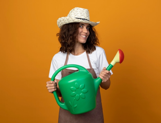 Smiling young female gardener in uniform wearing gardening hat holding watering can