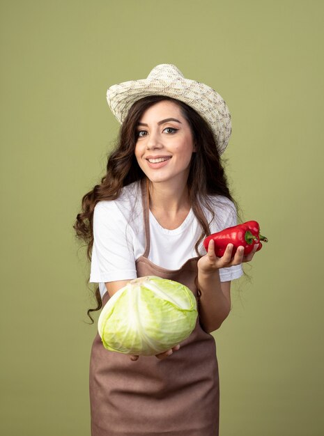 Smiling young female gardener in uniform wearing gardening hat holds cabbage and red peppers isolated on olive green wall