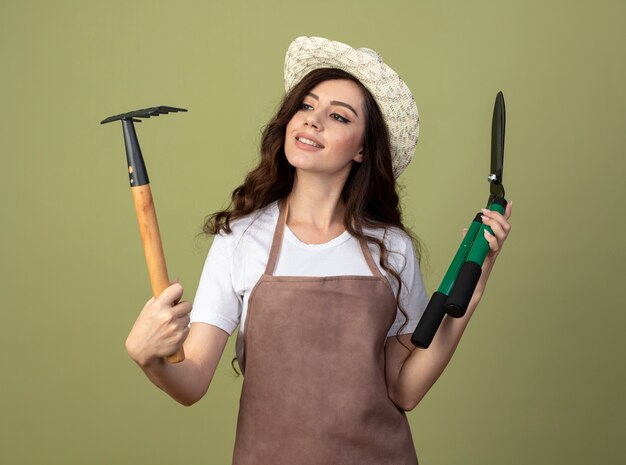 Smiling young female gardener in uniform wearing gardening hat holds clippers and looks at rake isolated on olive green wall