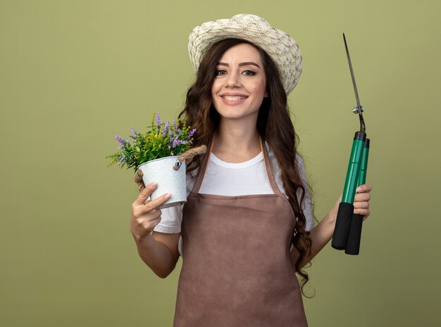 Smiling young female gardener in uniform wearing gardening hat holds flowerpot and garden clippers isolated on olive green wall with copy space