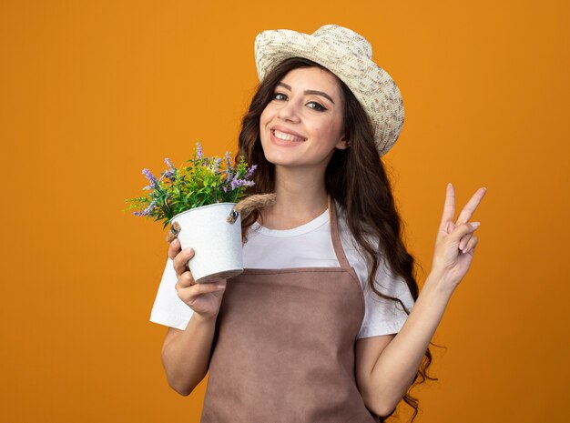 Smiling young female gardener in uniform wearing gardening hat holds flowerpot and gestures victory hand sign isolated on orange wall with copy space