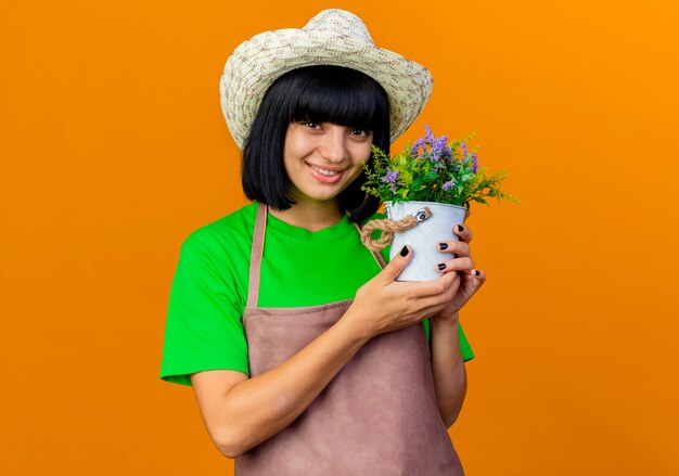 Улыбающаяся молодая женщина-садовник в униформе в садовой шляпе держит цветочный горшок