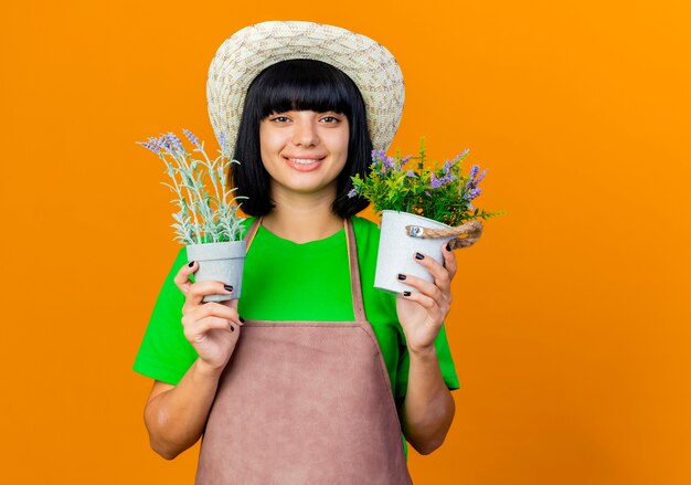 ガーデニング帽子をかぶって制服を着た若い女性の庭師の笑顔は植木鉢を保持します