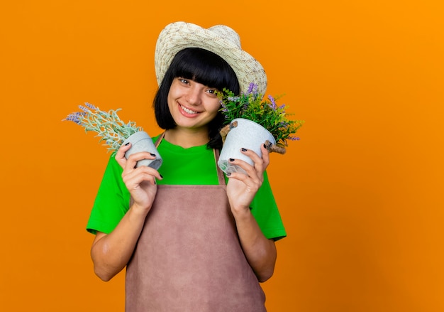 Smiling young female gardener in uniform wearing gardening hat holds flowerpots