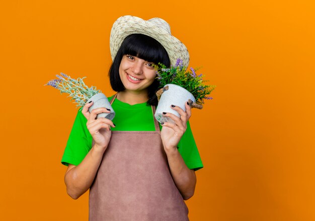 Smiling young female gardener in uniform wearing gardening hat holds flowerpots