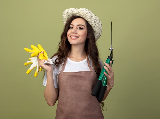 Smiling young female gardener in uniform wearing gardening hat holds garden clippers and gloves isolated on olive green wall