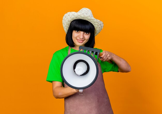 Foto gratuita sorridente giovane giardiniere femminile in uniforme che indossa un cappello da giardinaggio tiene un altoparlante