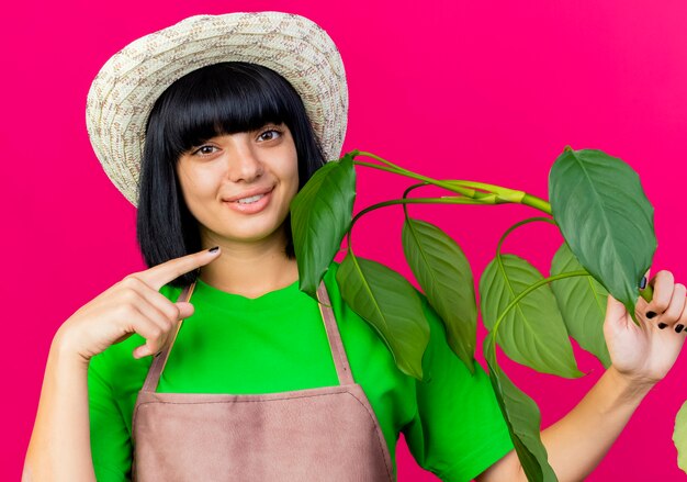 ガーデニング帽子をかぶって制服を着た若い女性の庭師の笑顔は、植物を保持し、ポイントします