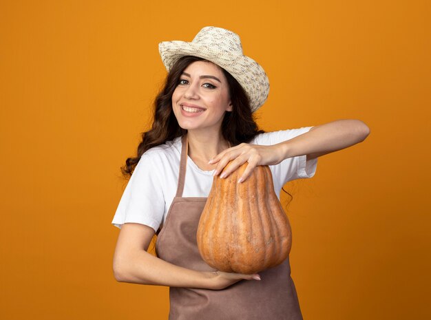 Smiling young female gardener in uniform wearing gardening hat holds pumpkin isolated on orange wall