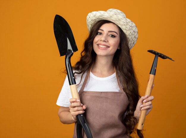 Улыбающаяся молодая женщина-садовник в униформе в садовой шляпе держит лопату и грабли, изолированные на оранжевой стене с копией пространства