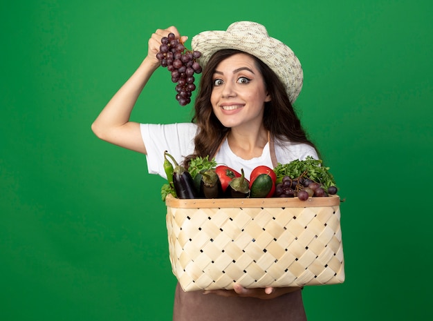 Sorridente giovane giardiniere femminile in uniforme che indossa il cappello da giardinaggio tiene cesto di verdure e uva isolato sulla parete verde