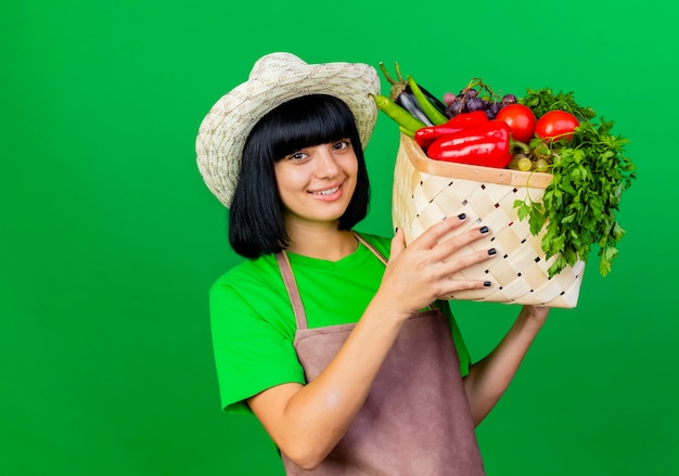 ガーデニング帽子をかぶって制服を着た若い女性の庭師の笑顔は野菜バスケットを保持