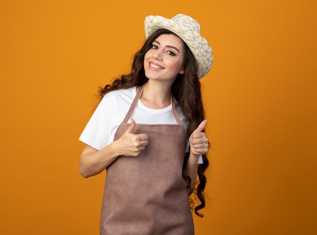 Smiling young female gardener in uniform wearing gardening hat thumbs up of two hands isolated on orange wall with copy space