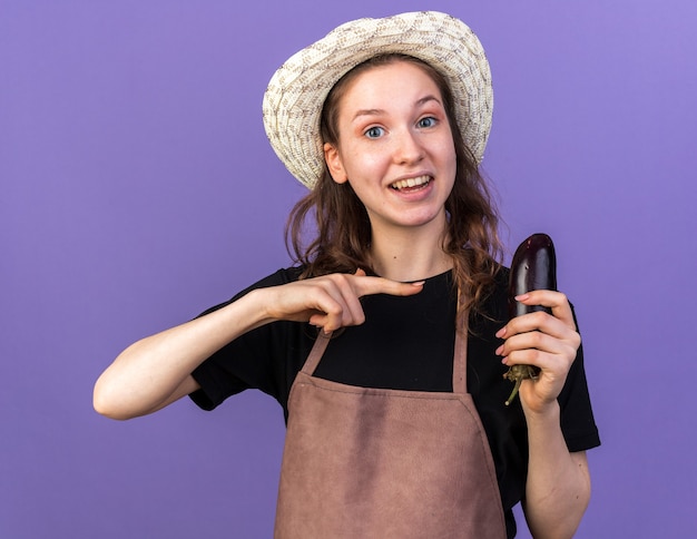 Smiling young female gardener wearing gardening hat holding and points at eggplant isolated on blue wall