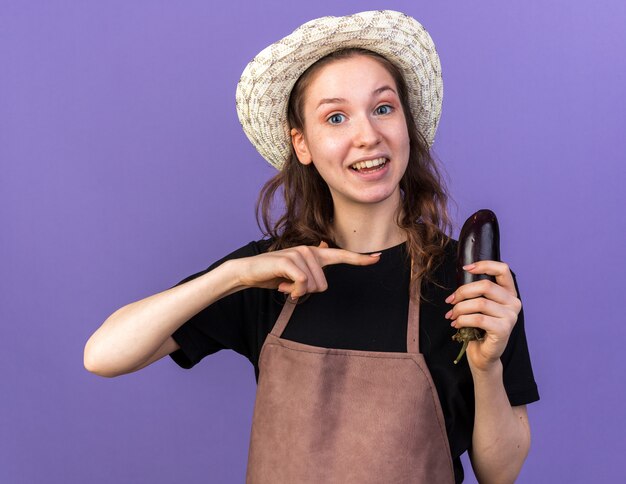Smiling young female gardener wearing gardening hat holding and points at eggplant isolated on blue wall