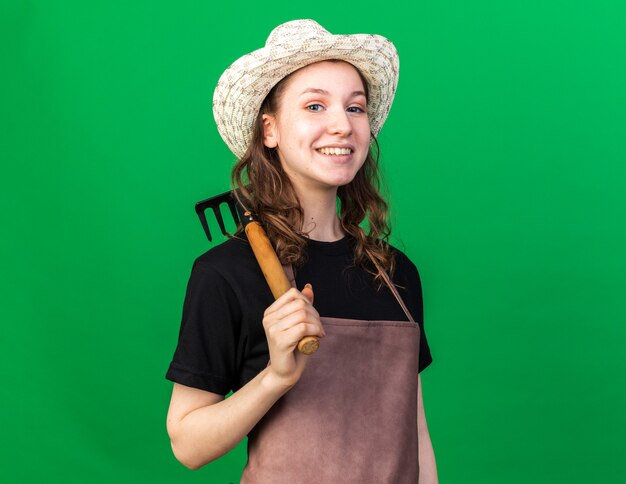 Smiling young female gardener wearing gardening hat holding rake on shoulder 