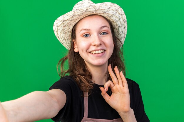 緑の壁に分離された大丈夫なジェスチャーを示すガーデニング帽子を身に着けている若い女性の庭師の笑顔