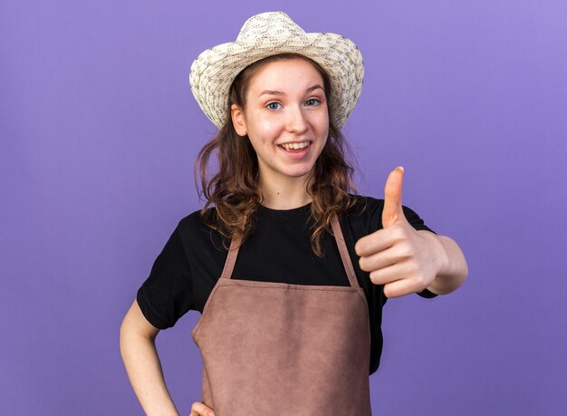 Smiling young female gardener wearing gardening hat showing thumb up isolated on blue wall