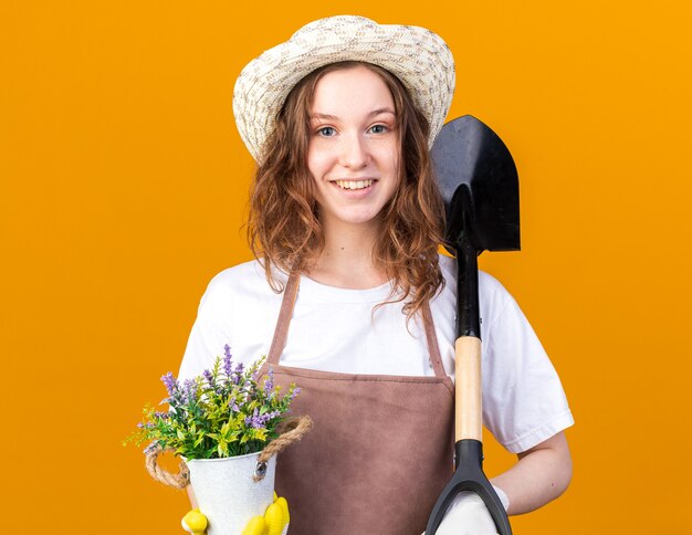 주황색 벽에 격리된 삽으로 화분에 꽃을 들고 장갑을 끼고 원예용 모자를 쓰고 웃고 있는 젊은 여성 정원사
