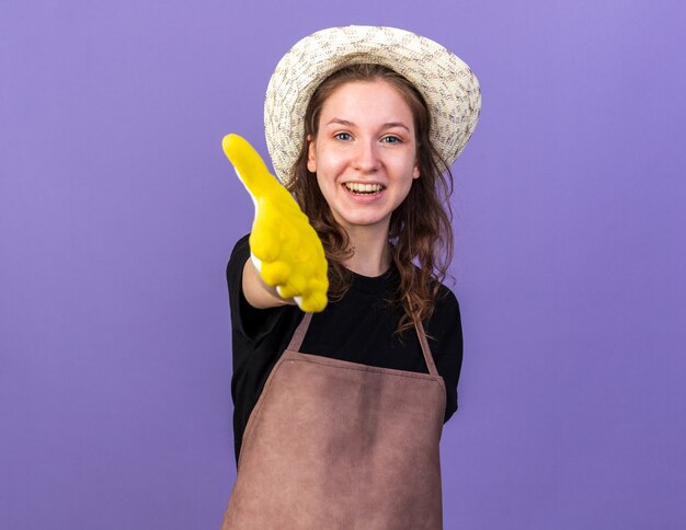 Улыбающаяся молодая женщина-садовник в садовой шляпе с перчатками, протягивая руку, изолированную на синей стене