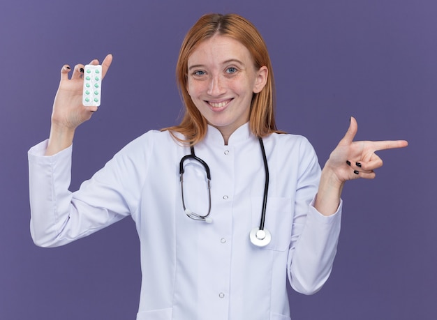無料写真 紫色の壁に隔離された側を指しているカメラに医療薬のパックを示す医療ローブと聴診器を身に着けている若い女性生姜医師の笑顔