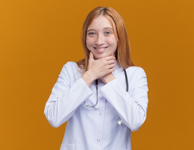あごに手を置いて医療ローブと聴診器を身に着けている若い女性の生姜医師の笑顔