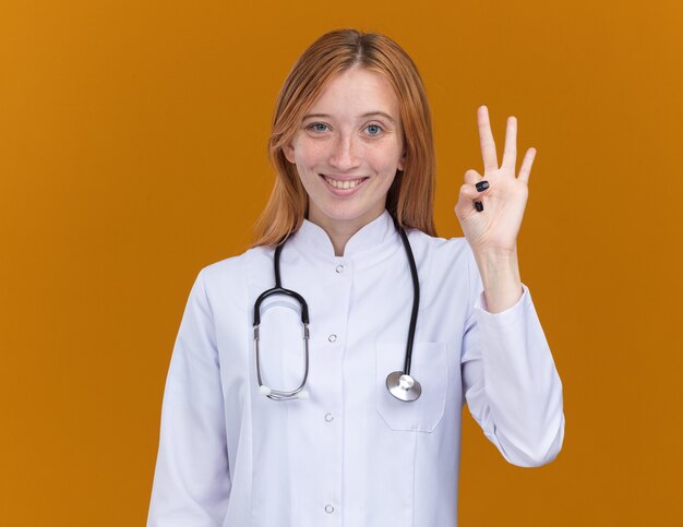 Smiling young female ginger doctor wearing medical robe and stethoscope looking at front doing ok sign isolated on orange wall