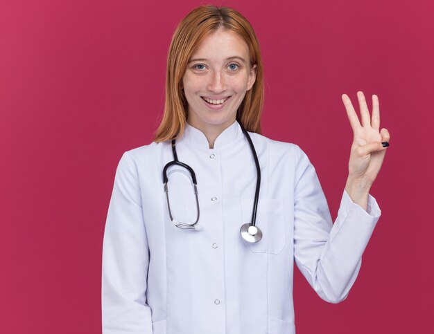 真っ赤な壁に隔離された手で3つを示す正面を見て医療ローブと聴診器を身に着けている若い女性生姜医師の笑顔
