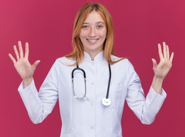 真っ赤な壁に隔離された空の手を示す医療ローブと聴診器を身に着けている若い女性生姜医師の笑顔