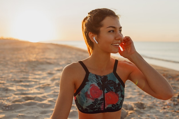 Smiling young female listening music in headphones after training. Young athletic woman exercising near the sea . Summer sunset.
