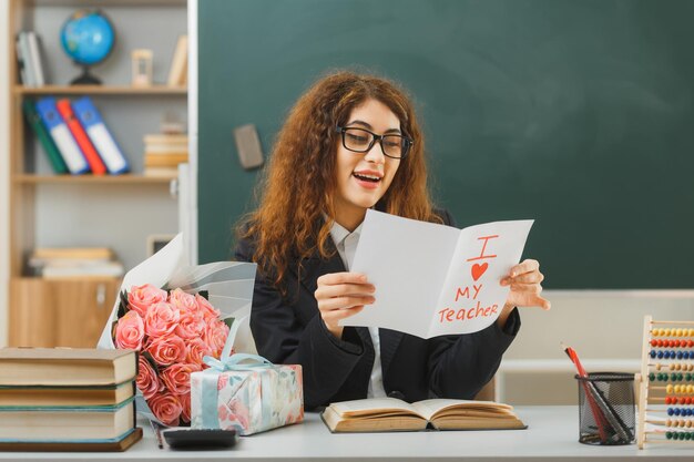 smiling young female teacher holding and looking at postcard sitting at desk with school tools in classroom