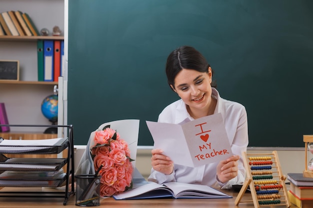 Sorridente giovane insegnante femminile che tiene e guarda la cartolina seduta alla scrivania con gli strumenti della scuola in classe