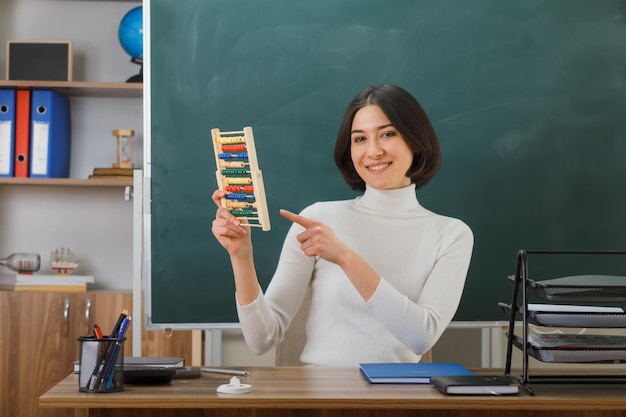 Free photo smiling young female teacher holding and points at abacus sitting at desk with school tools on in classroom