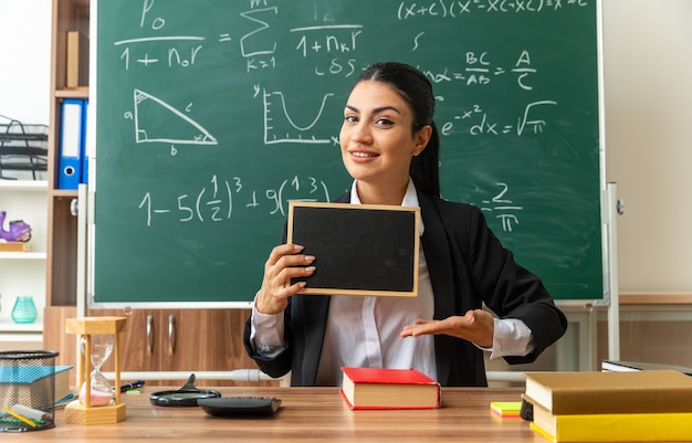smiling young female teacher sits at table with school supplies holding and points at mini blackboard in classroom