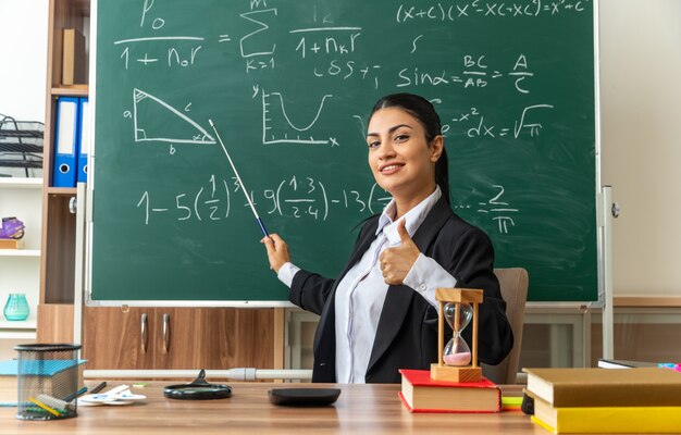 улыбается молодая учительница сидит за столом со школьными принадлежностями указывает на доску с указателем, показывая большой палец вверх в классе