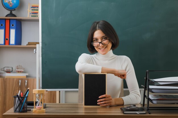 smiling young female teacher wearing glasses holding mini chalkboard sitting at desk with school tools on in classroom